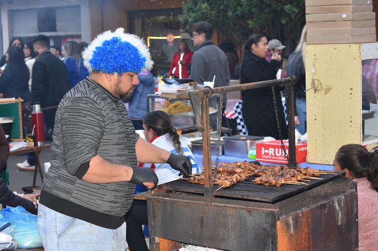Hombre con cabello rizado azul y blanco, usando guantes negros, cocina brochetas de carne mientras un grupo observa en un ambiente festivo.