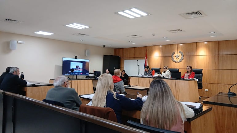 Mujer con blazer rojo y cuatro personas en el panel, mientras otros asistentes toman notas en sala judicial.