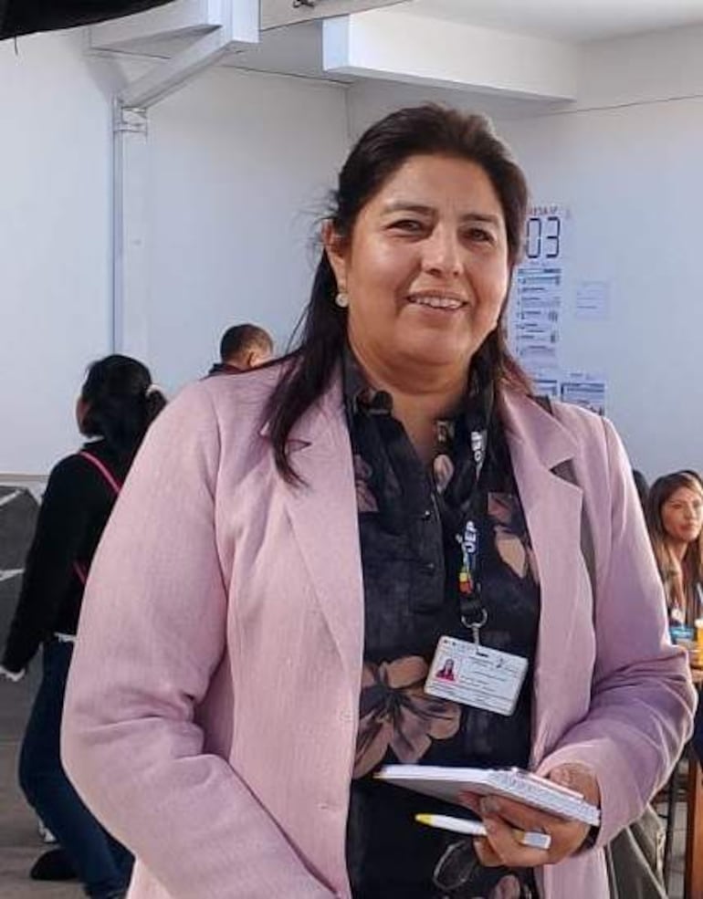 Mujer con blusa negra y blazer rosa sonríe, sosteniendo una libreta en un ambiente iluminado con personas al fondo.