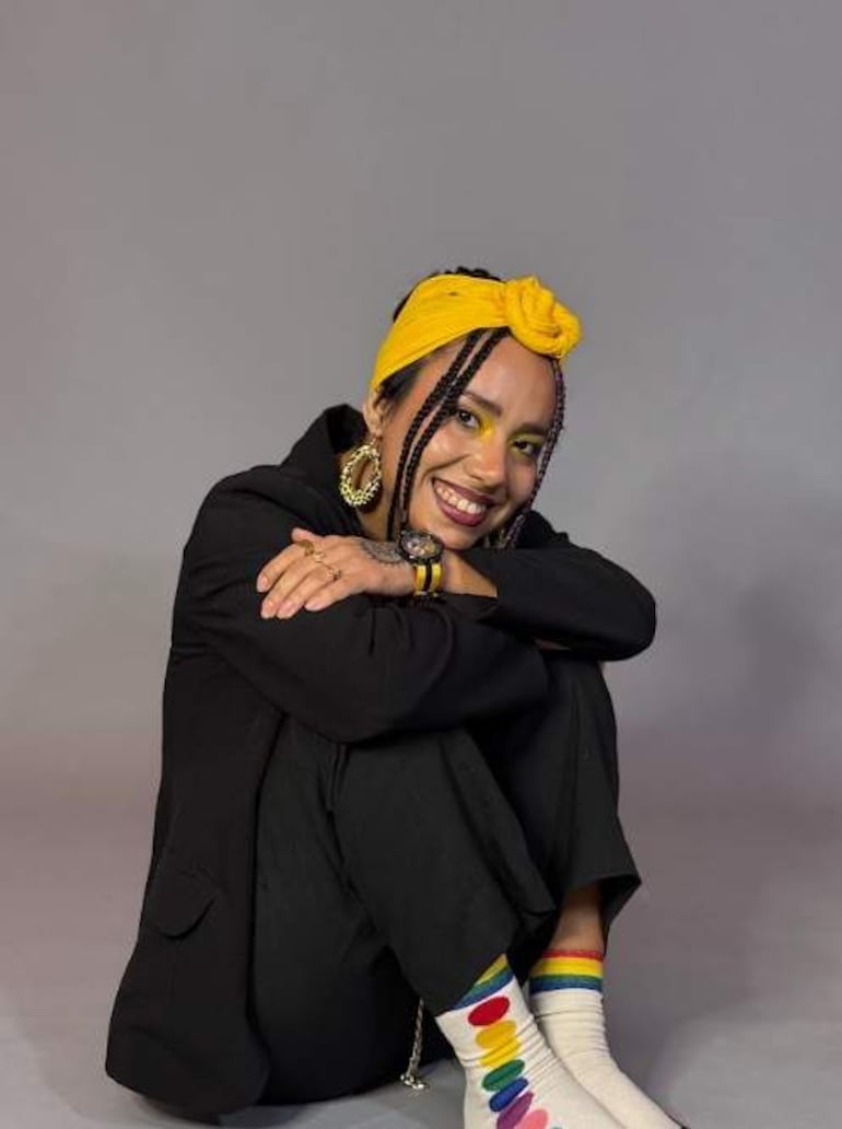 Mujer con pañuelo amarillo en la cabeza y atuendo oscuro, sonriendo en un entorno de estudio con decoración colorida.
