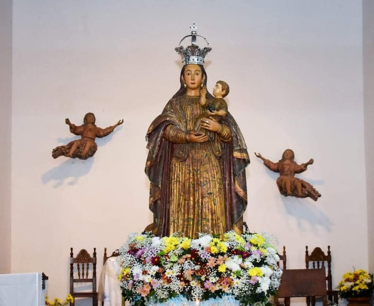 Altar con la figura de la Virgen María sosteniendo al Niño Jesús y decorado con flores de colores, en ambiente religioso solemnemente iluminado.