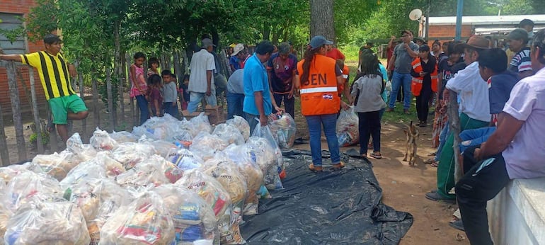 Grupo de personas en área rural, mujer con chaleco naranja 'SEN' en el centro, distribuyendo comida sobre lona negra.