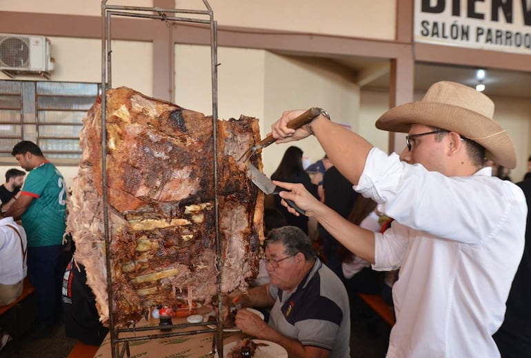 Hombre con sombrero de cowboy y camisa blanca corta carne asada, mientras un comensal con camiseta verde disfruta de su comida.