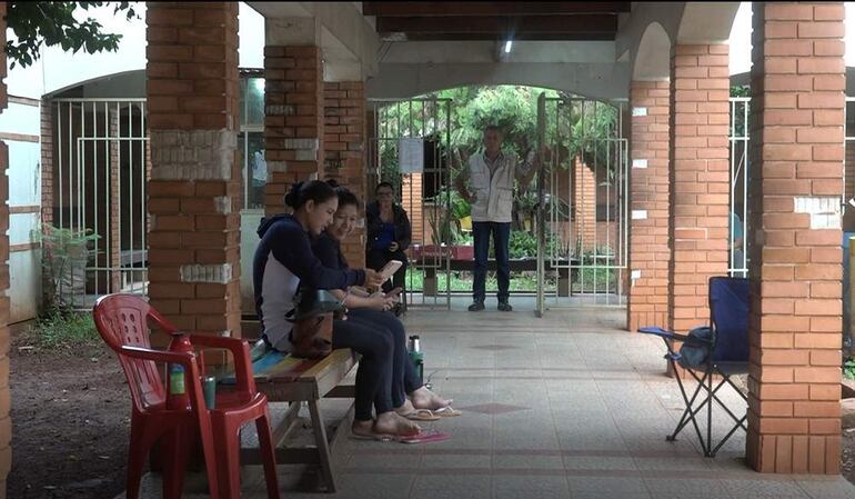 Dos mujeres sentadas en un banco con teléfonos; otra mujer de pie observa. Entorno tranquilo con sillas plásticas.