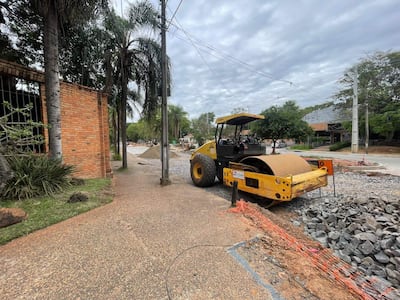 Rodillo compactador amarillo estacionado en grava, con pared de ladrillo y palmeras al lado, bajo un cielo nublado.