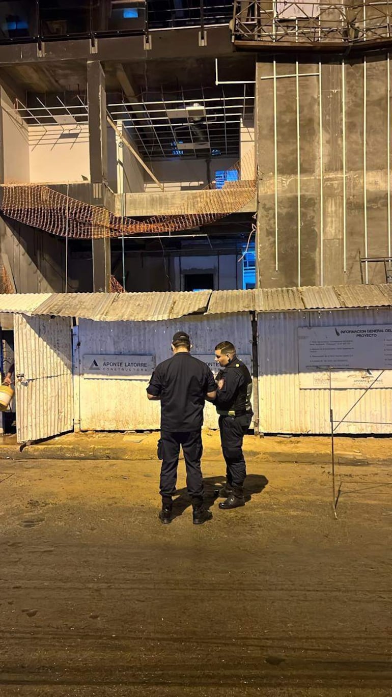 Hombres en uniformes oscuros revisan documentos frente a una obra en construcción, con andamios y mallas de seguridad visibles.