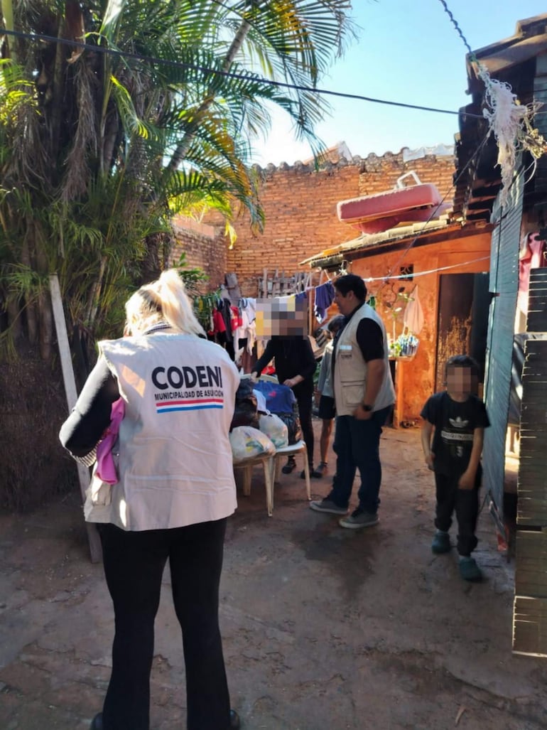 Mujer con chaleco del CODENI sostiene un objeto y habla con un hombre en un barrio soleado, rodeados de adultos y niños.