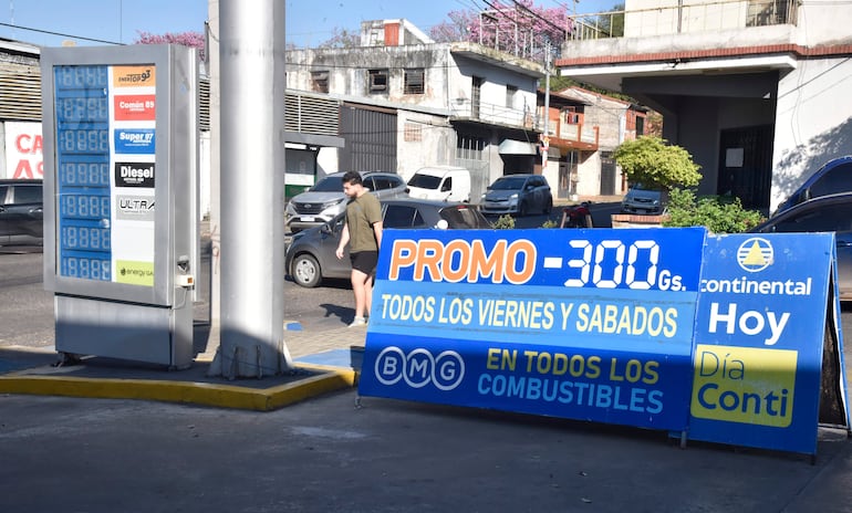 Persona en pantalón corto y camiseta verde camina hacia cartelera de promoción de combustibles en estación de servicio.