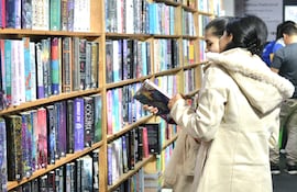 Mujer con abrigo claro en estante de libros, observando atentamente varias publicaciones en un ambiente de feria literaria.