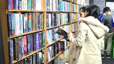 Mujer con abrigo claro en estante de libros, observando atentamente varias publicaciones en un ambiente de feria literaria.