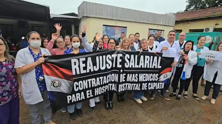 Grupo de médicos en uniforme sosteniendo pancarta que dice 'REAJUSTE SALARIAL' durante la protesta frente al hospital.