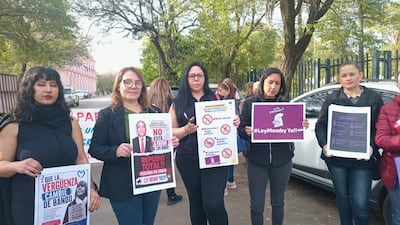 Seis mujeres, algunas con carteles, protestan con expresiones decididas en un espacio urbano.