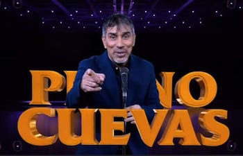 Nelson Cuevas Pipino sonríe y gesticula mientras habla frente a un micrófono, vestido con un traje oscuro en un escenario iluminado.