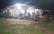Grupo de personas bajo techo en ambiente rural, algunas sentadas y otras de pie, en reunión social con sillas y mesa.