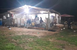 Grupo de personas bajo techo en ambiente rural, algunas sentadas y otras de pie, en reunión social con sillas y mesa.