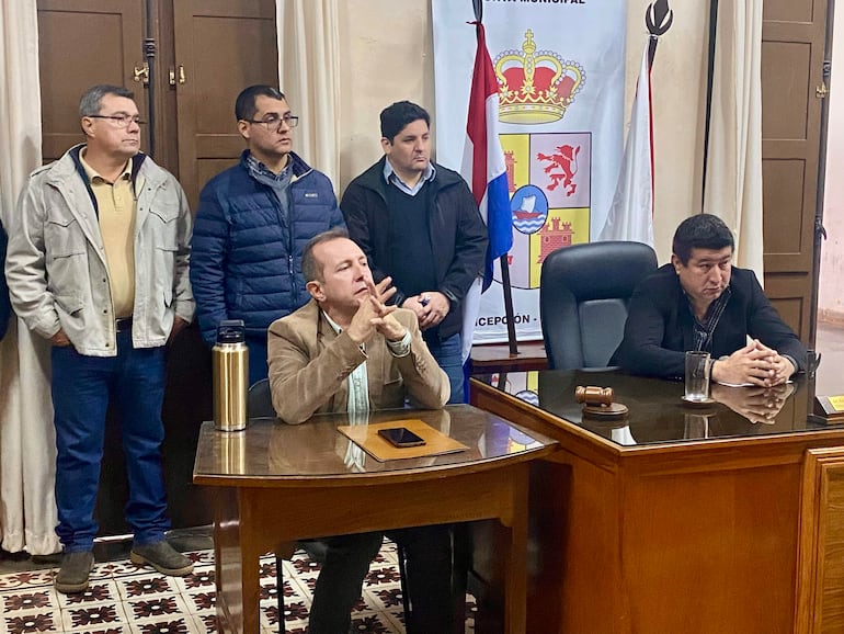 Cinco hombres en sala municipal, uno sentado pensativo y cuatro de pie escuchando atentamente, con vaso y martillo sobre la mesa.