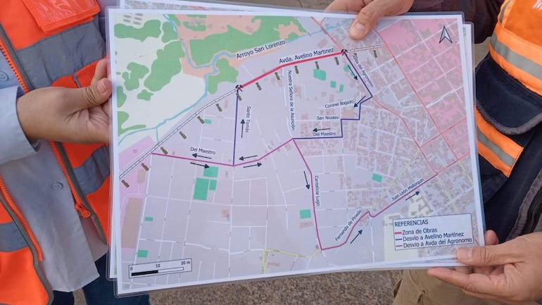 Dos personas, una en chaleco naranja, sostienen un mapa en un entorno urbano con suelo de asfalto, centrados en la discusión de proyectos.