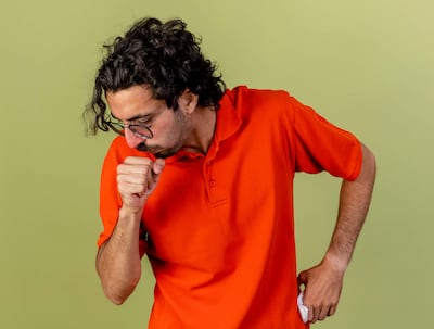 Hombre con camiseta naranja tosiendo en un ambiente interior de fondo verde, sin otras personas presentes.