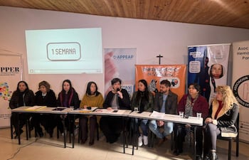 Mujer con micrófono habla en mesa rodeada de asistentes atentos. Carteles sobre enfermedades raras en el fondo.