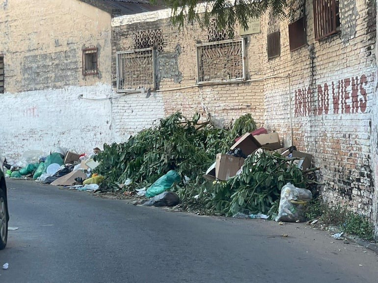 Ambiente urbano con acumulación de basura variada en calles de Asunción. Pared blanca deteriorada y graffiti visible.