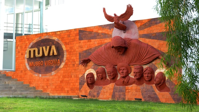 Mural en el MUVA con esculturas de rostros y un águila, sobre fondo naranjado que incluye el texto 'Museo Viedma'.