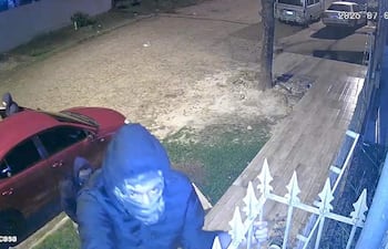 Un hombre con capucha intenta escalar una reja mientras otro asiste cerca de un automóvil rojo en una escena nocturna.