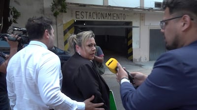 Zenaida Delgado con cabello rubio recogido, seria, mientras un hombre en traje oscuro le sostiene un micrófono amarillo en un garaje.