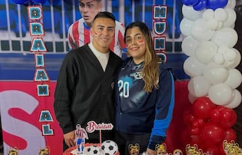 Braian, con camiseta blanca y abrigo negro, sonríe junto a una amiga en chaqueta azul, rodeados de pastel decorado y globos.