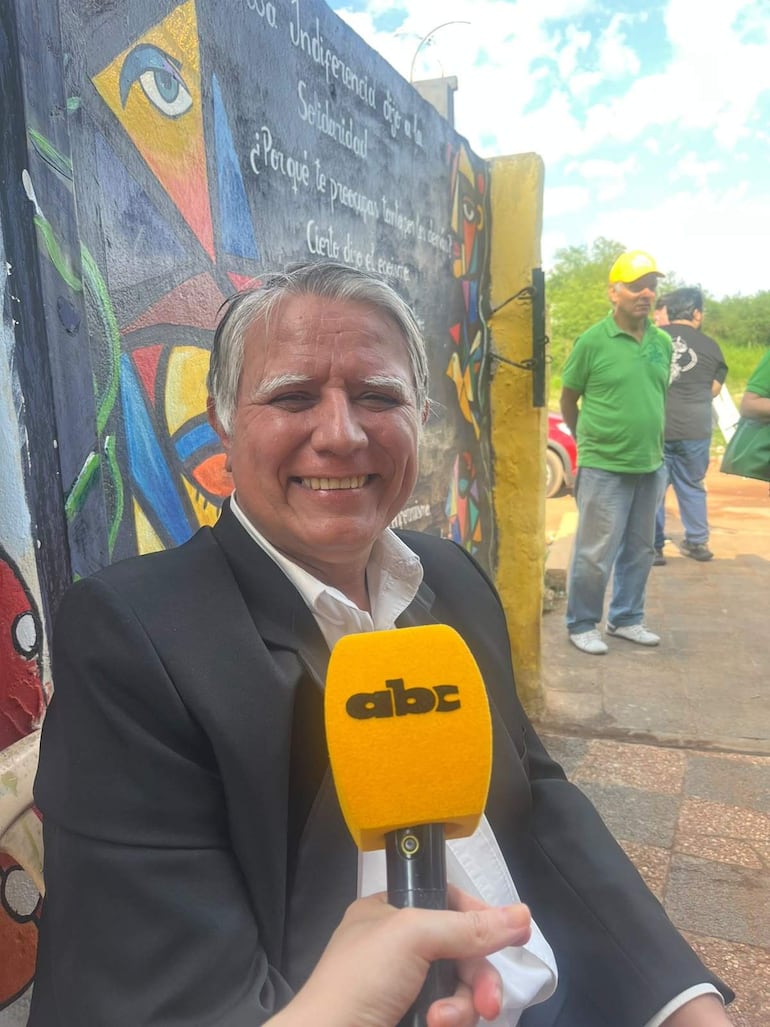 Hombre con chaqueta negra y camisa blanca sonríe mientras habla a un micrófono amarillo en evento comunitario.