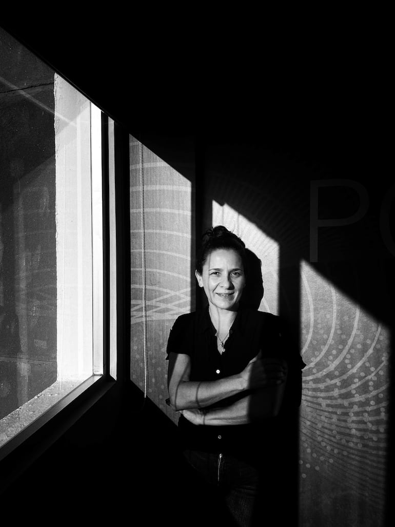 Mujer con camisa oscura y jeans, de pie junto a una ventana, con una expresión serena en un ambiente interior en blanco y negro.