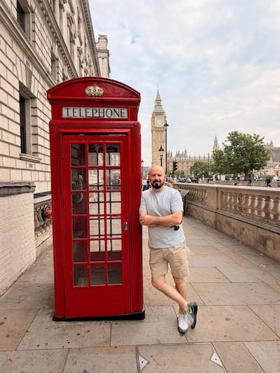 Hombre con camiseta gris y pantalones cortos claros junto a cabina telefónica roja, con clima nublado y Big Ben detrás.