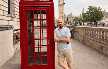 Hombre con camiseta gris y pantalones cortos claros junto a cabina telefónica roja, con clima nublado y Big Ben detrás.