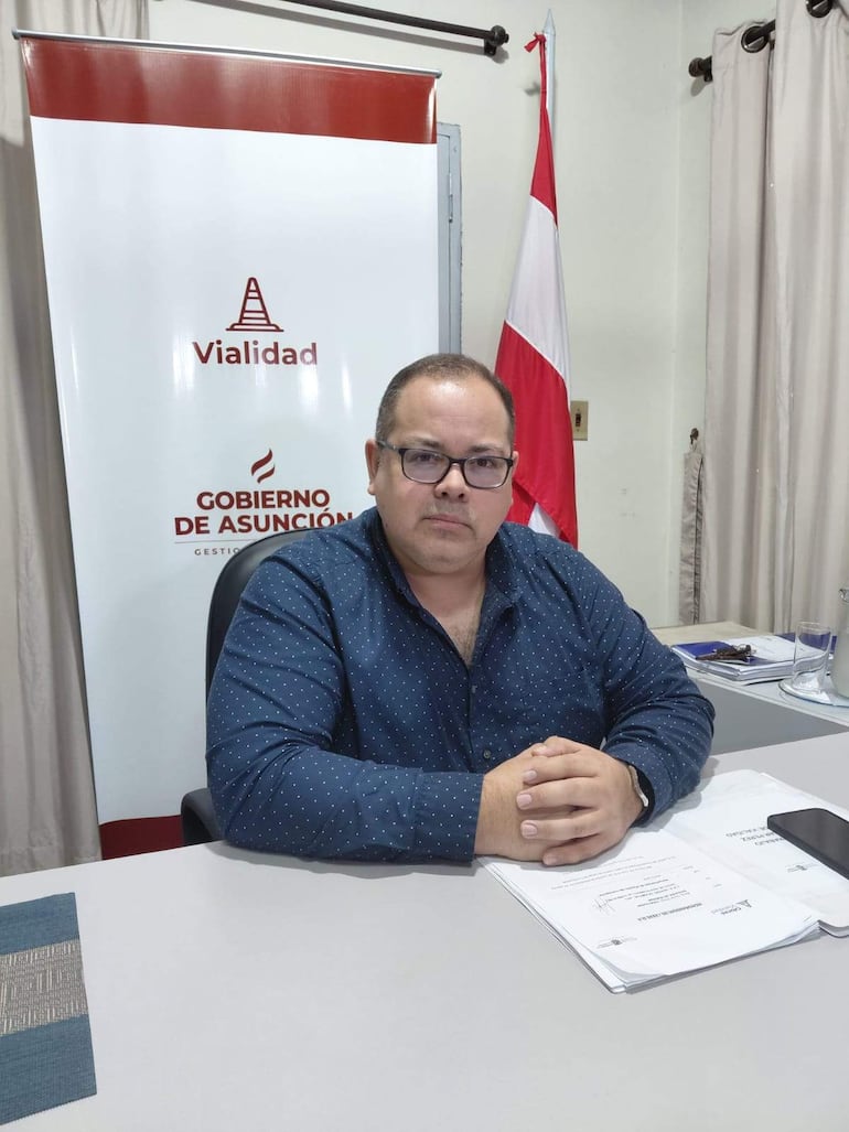 Óscar Pérez, con camisa azul con puntos, revisa documentos en su oficina, con estandarte del Gobierno de Asunción al fondo.