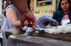 Una mano masculina alza un frasco de tinta, mientras dos mujeres revisan documentos en un centro de votación.