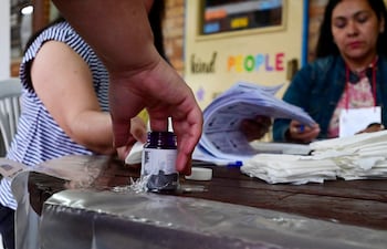 Una mano masculina alza un frasco de tinta, mientras dos mujeres revisan documentos en un centro de votación.