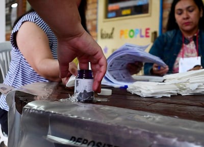 Una mano masculina alza un frasco de tinta, mientras dos mujeres revisan documentos en un centro de votación.