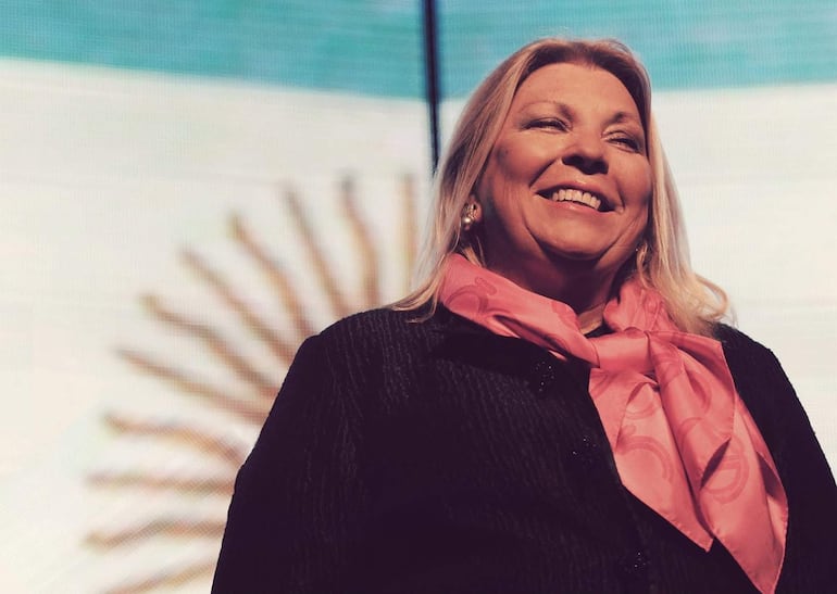 Elisa Carrió sonriendo, con cabello rubio suelto y vestimenta elegante, iluminada por un ambiente festivo.