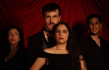 Cuatro miembros del elenco, dos mujeres con vestidos oscuros y dos hombres con atuendos serios, en un fondo de tela roja.