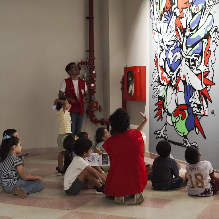 Niños sentados con camisetas coloridas, observan a un adulto de espaldas con camiseta roja, frente a un mural vibrante.