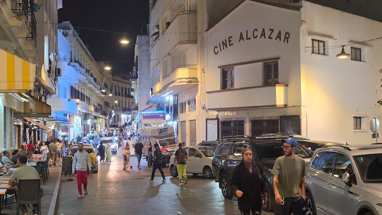 Calle iluminada de Tánger con personas caminando, sentadas en mesas afuera y el cine Alcazar visible al fondo.