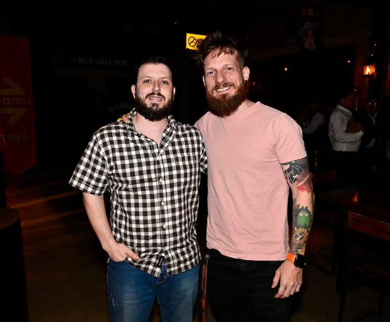 Dos hombres, uno con camisa de cuadros y el otro con camiseta rosa, sonriendo en un ambiente acogedor de bar con luces tenues.