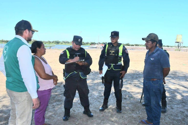Policía toma notas mientras un hombre de chaleco verde y una mujer observan en un entorno rural con árboles y un depósito de agua.
