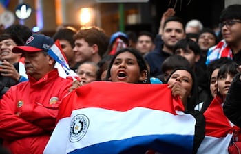 Joven con bandera de Paraguay y gorra roja, rodeada de personas alegres en un evento festivo al aire libre.