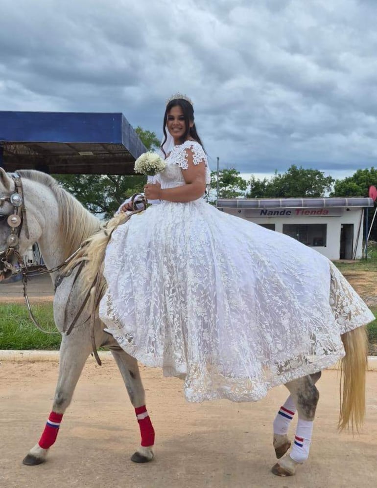 Mujer en vestido blanco y tiara sonriente, montando un caballo decorado con campanas, en un entorno rural.