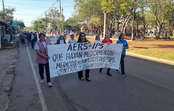 Mujeres con camisetas de salud sostienen pancarta sobre medicamentos y especialistas, marchando en un entorno urbano.