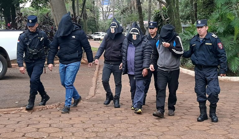 Cinco hombres con capuchas son escoltados por tres policías uniformados en un entorno sereno y arbolado.