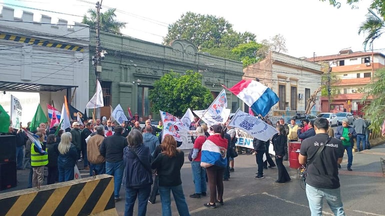 Grupo de manifestantes sosteniendo banderas y pancartas en un entorno urbano con edificios clásicos de fondo.