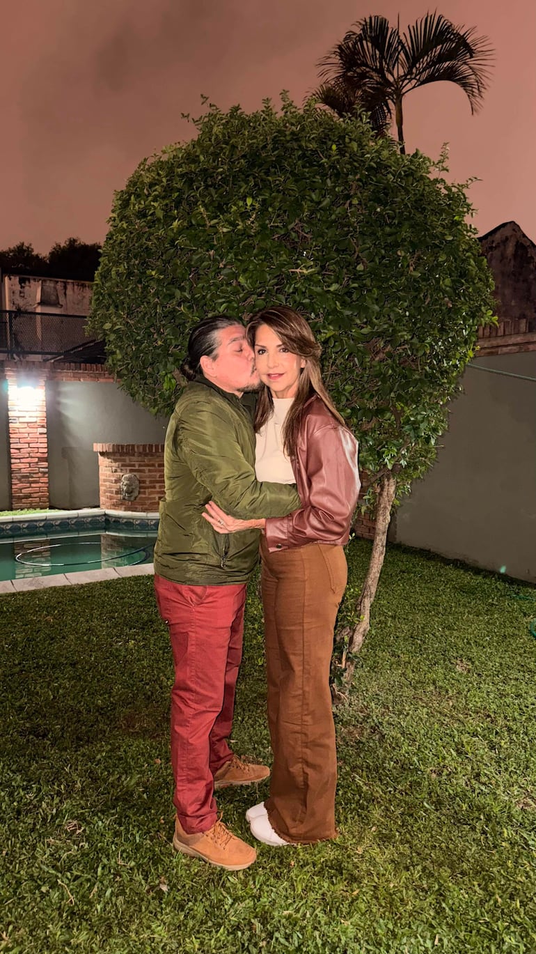 Hombre con chaqueta verde y pantalones rojos, sonríe mientras se inclina hacia una mujer en chaqueta marrón y pantalones claros.