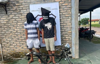 Dos hombres con rostros cubiertos, uno en camiseta negra y otro en camiseta a rayas, en un ambiente policial con piezas de motocicleta detrás.