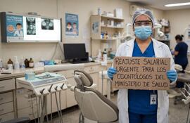 Odontólogo con bata blanca y guantes sostiene un cartel en protesta, fondo de consultorio dental y equipo médico visible.